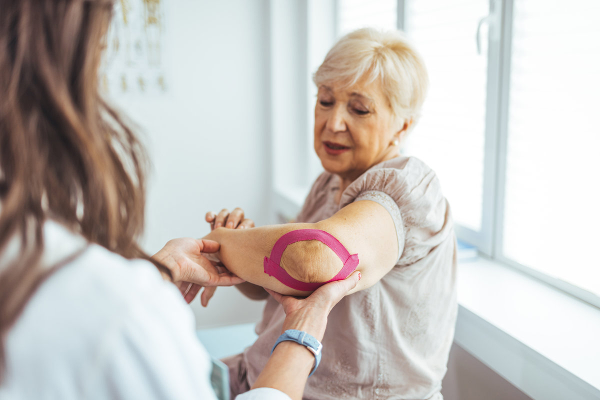 Kinesiology taping treatment with pink tape on senior patient injured arm. Woman hands apply kinesio treatment after muscle injury. A Modern rehabilitation physiotherapy worker with woman client